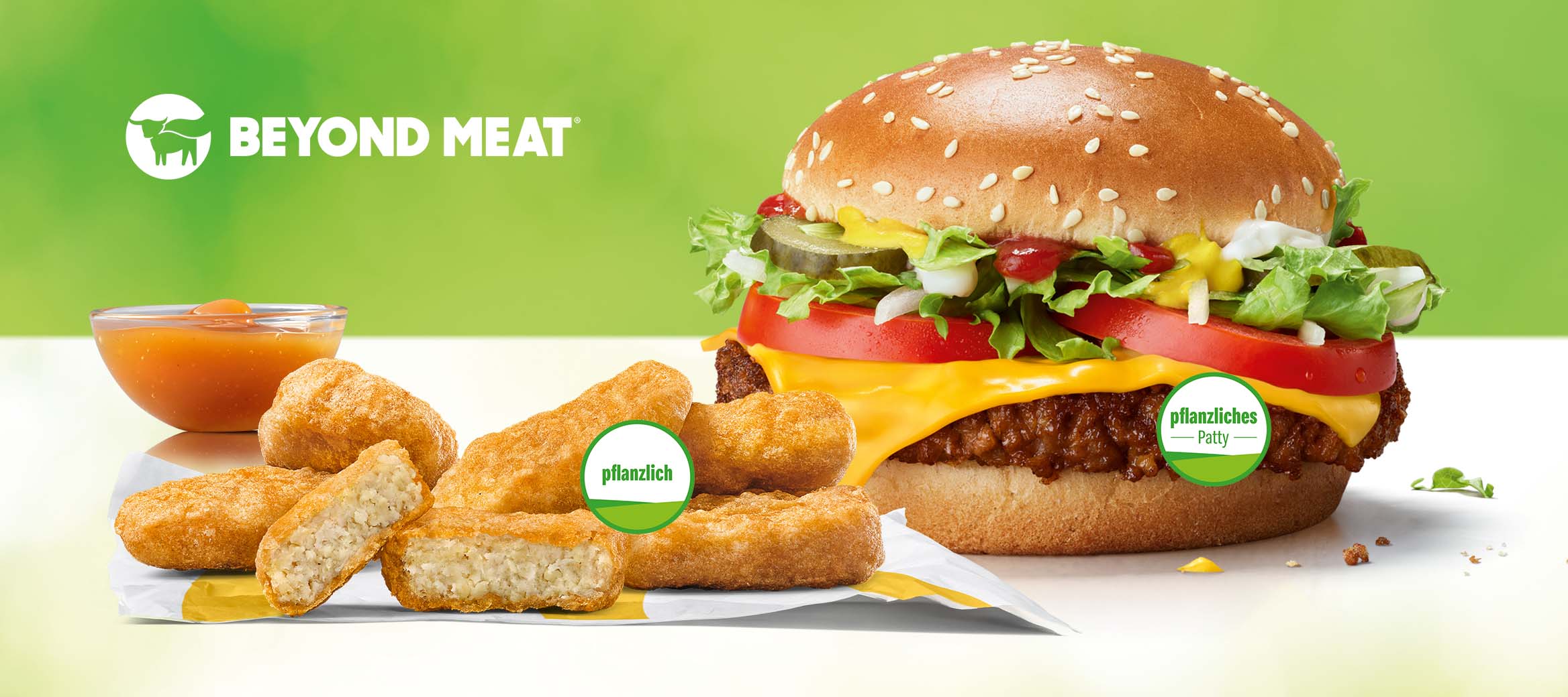 Ein McPlant® auf dem “pflanziliches Patty“ steht. Daneben McPlant® Nuggets, auf denen „pflanzlich“ steht vor einem Schälchen Süß-sauer Sauce. Darüber steht Beyond Meat.