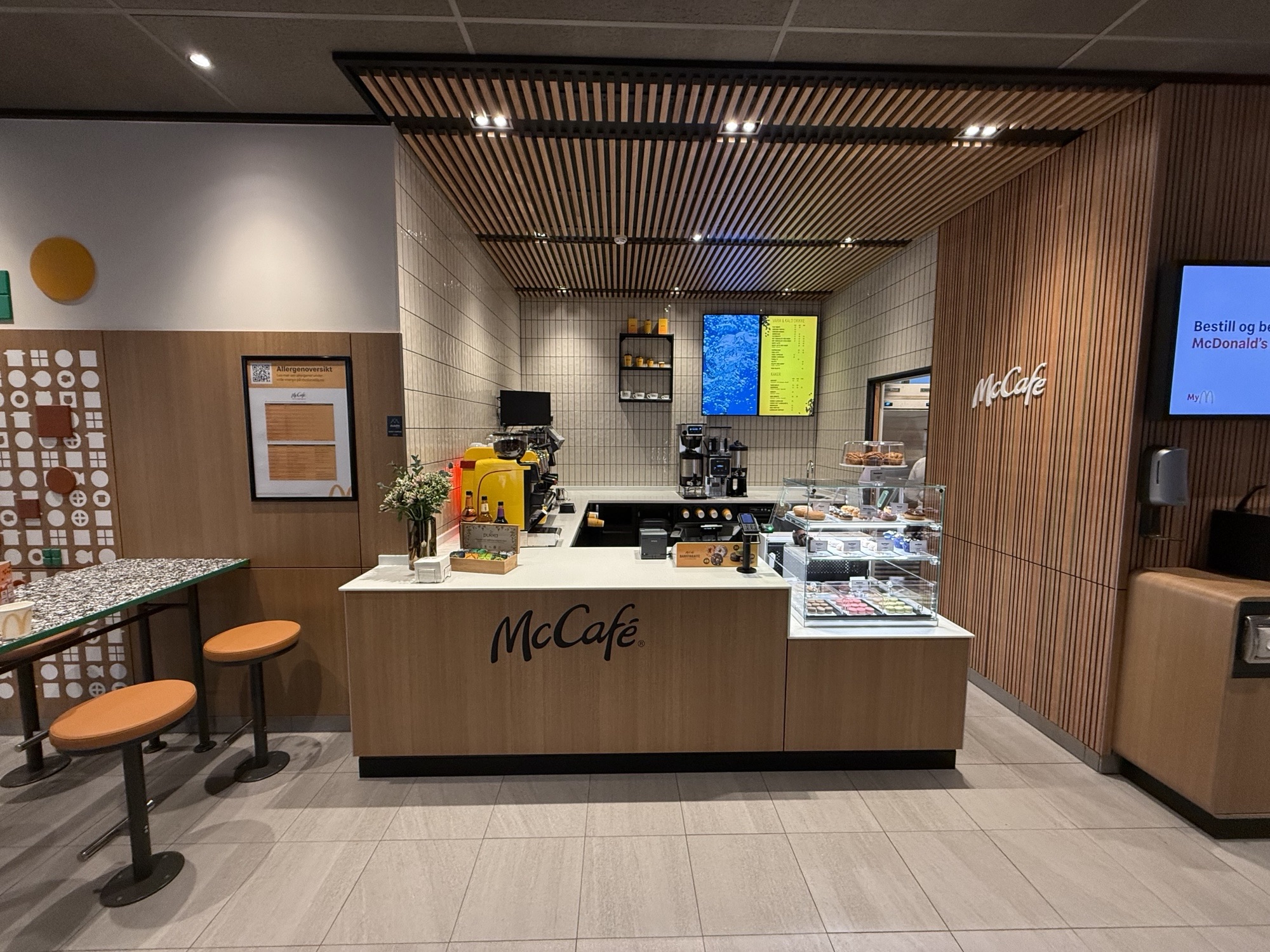  Interiør av en McCafé-kafé inne i en McDonald's-restaurant. Bildet viser en minimalistisk kaffebar med trepanelvegger og et hvitt flislagt bakgrunnsområde. Det er en disk med kaffemaskiner, kake- og bakevarer i en glassmonter, samt en digital meny på veggen. Ved siden av disken er det høye sitteplasser med oransje stoler og et bord med et flekkete design. Rommet har et moderne og stilrent design med et tak laget av trelister som skaper en varm atmosfære. Foto.