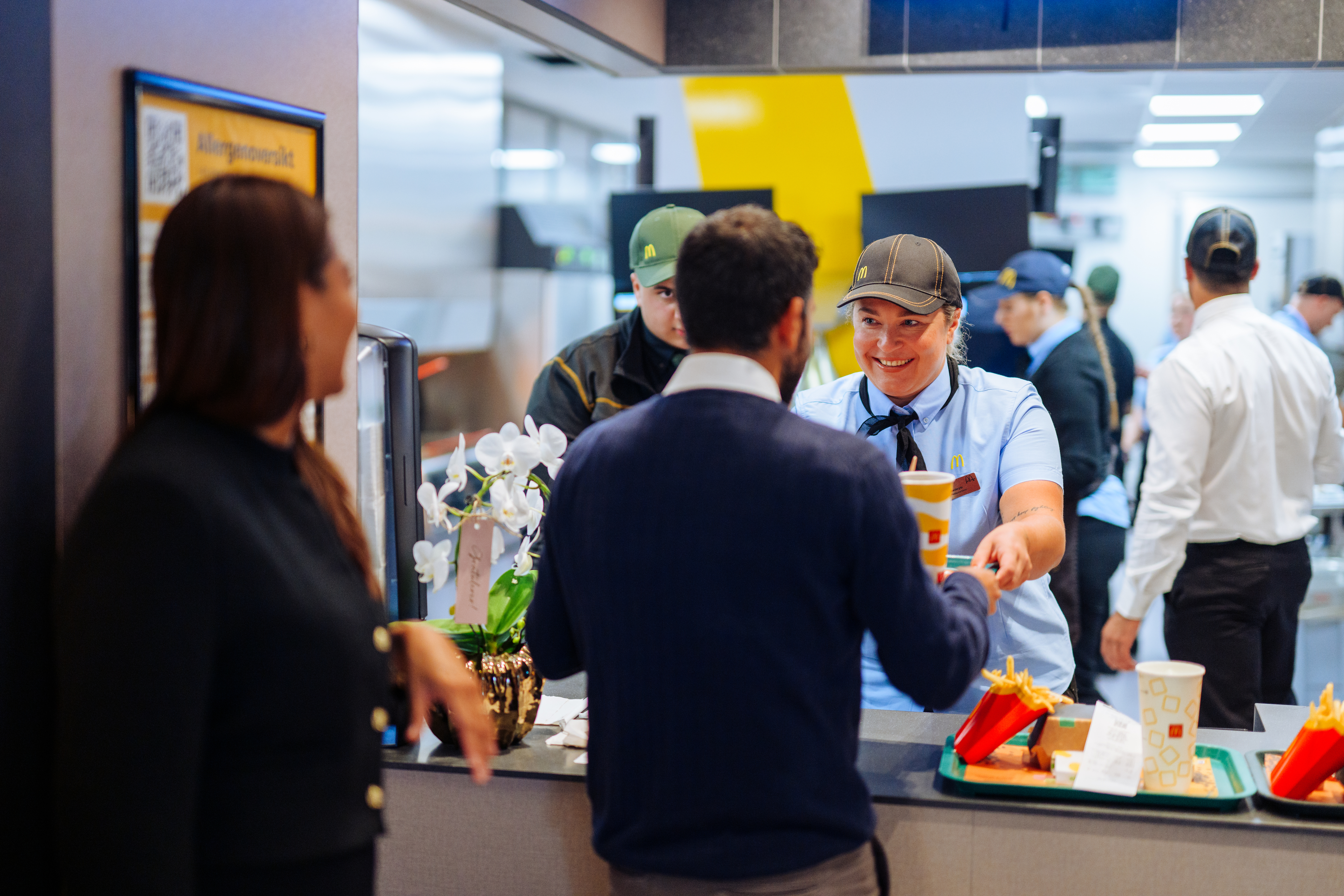 En McDonald's-ansatt i uniform smiler mens hun overrekker en drikke til en kunde ved kassen. Andre kunder og ansatte er synlige i bakgrunnen, og et brett med pommes frites står på disken.